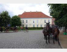 Pflasterstraße mit einer Pferdekutsche im Vordergrund und zwei Pferden, die rechts im Bild stehen. Im Hintergrund ein gelbes, zweigeschossiges Gebäude mit roten Ziegeldach, umgeben von Bäumen und Fahrradfahrern.