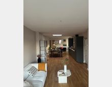 Moderne Wohnungsinneneinrichtung mit offenem Raumkonzept, bestehend aus einem Wohnzimmer mit einem weißen Sofa und Couchtisch im Vordergrund, einem Essbereich mit hölzernem Esstisch und Stühlen in der Mitte sowie einer offenen Küche mit dunklen Schränken im Hintergrund. Heller Holzboden und minimalistische Dekoration prägen den Raum.