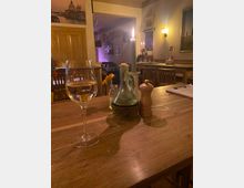 Innenraum eines gemütlichen Restaurants mit Holztischen und -stühlen, warmen Wandlampen und Bildern an den Wänden. Im Vordergrund steht auf einem Holztisch ein Weinglas sowie Kerze, Glasflasche, kleine Blumenvase und Gewürzmühle.