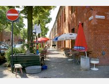 Straßenszene in einem Außenbereich mit Kopfsteinpflaster, flankiert von roten Backsteingebäuden. Links stehen parkende Autos und ein Baum, während rechts Tische und Stühle unter Sonnenschirmen vor einem Restaurant aufgestellt sind; ein „Einfahrt verboten“-Schild ist sichtbar.