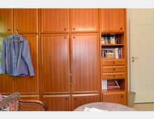 Holzvertäfelter Kleiderschrank mit mehreren Schubladen und Einlegefächern auf der rechten Seite, an einer Wand in einem Wohnraum. Links hängt ein blaues Hemd an einem Kleiderhaken, und im Vordergrund ist ein runder Tisch teilweise sichtbar.