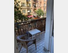 Ein kleiner Balkon mit gefliestem Boden, einem weißen Tisch, einem geflochtenen Stuhl und einem Sichtschutz aus Holzgeflecht. Im Hintergrund sind bunte Altbauten mit Balkonen, ein Baum und Geschäfte, darunter ein Spätkauf, zu sehen.