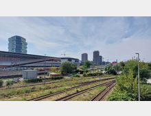 Bahnhofsumgebung mit mehreren Gleisen im Vordergrund, umgeben von Vegetation. Im Hintergrund sind ein modernes Einkaufszentrum mit der Aufschrift „Riverside Mall“, ein moderner Bürogebäudeturm sowie zwei im Bau befindliche Hochhäuser zu sehen, zusammen mit Baukränen und weiteren städtischen Elementen.