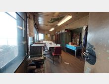 Ein einfacher Arbeitsraum mit abgenutztem Holzboden, einer langen Tabelle in der Mitte, umgeben von Stühlen. An den Wänden befinden sich Metallspinde, ein Fenster mit Blick nach draußen und eine Pinnwand mit Aufklebern; die Decke zeigt abgeblätterte Farbe.