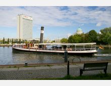 Ein historisches Dampfschiff mit der Aufschrift „Havel Dampfschifffahrt Potsdam“ fährt auf einem Fluss, im Hintergrund ist ein modernes Hochhaus sowie die grüne Kuppel einer Kirche zu sehen. Im Vordergrund befindet sich eine Uferpromenade mit einer Parkbank und Kopfsteinpflaster.