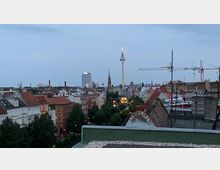 Stadtpanorama von Berlin bei Dämmerung mit Blick auf den Fernsehturm, umgeben von historischen Gebäuden mit roten Ziegeldächern und moderner Architektur. Im Vordergrund sind begrünte Straßenzüge und eine Dachlandschaft, im Hintergrund Baukräne sichtbar.