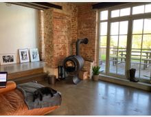 Wohnzimmer mit industriellem Stil und Ziegelwänden, großen Fenstern mit Blick auf eine Terrasse und Garten. Im Raum befinden sich ein schwarzer Holzofen, eingerahmte Schwarz-Weiß-Bilder, eine braune Ledercouch und ein schlafender schwarzer Katze.