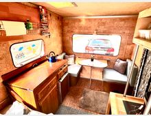 Innenansicht eines umgebauten Wohnwagens mit Holzverkleidung. Zu sehen sind eine kleine Küchenzeile mit blauem Wasserkocher, eine Sitzecke mit Tisch und Polstermöbeln sowie große Fenster, durch die ein geparktes Auto sichtbar ist.