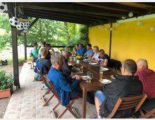 Eine Gruppe von Menschen sitzt unter einer überdachten Terrasse mit Holzbalken an einem langen Holztisch. Die Terrasse hat eine gelbe Wand im Hintergrund, dekorative Lampions und umgebende grüne Vegetation.