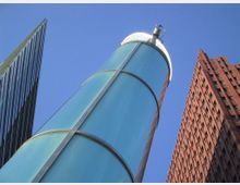 Ansicht moderner Architektur von unten mit drei Gebäuden: ein schlanker, verglaster Zylinder in der Mitte, ein Hochhaus mit Glasfassade links und ein rotes Gebäude mit Ziegeloptik rechts. Der Himmel ist klar und blau.