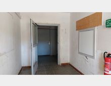 Ein schmaler Eingangsbereich mit weißen Wänden, abgenutztem Bodenbelag aus grauem Stein und einer geöffneten Glastür, die zu einer geschlossenen Metalltür im Hintergrund führt. An der rechten Wand befindet sich ein Feuerlöscher, ein leeres Wandfeld und ein Erste-Hilfe-Schild.