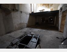 Innenraum eines verfallenen Industriegebäudes mit hohen, abblätternden Wänden, einer Zwischenebene und Licht, das durch Öffnungen im oberen Bereich einfällt. Auf dem staubigen Betonboden befindet sich eine große rechteckige Bodenöffnung mit Metallrahmen, im Hintergrund sind dunkle Türen oder Fensteröffnungen zu sehen.