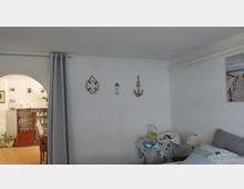 Heller Wohnraum mit weißen Wänden, maritimer Wanddekoration und einem Bett im Vordergrund; daneben stehen ein Babyhochstuhl und ein Nachttisch mit Lampe. Links führt ein Rundbogen in einen angrenzenden Essbereich mit Vitrine und Stühlen, getrennt durch einen grauen Vorhang.