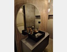 Modernes Badezimmer mit einem runden Wandspiegel, einer schwarzen Waschtischarmatur und einer minimalistisch gestalteten Waschbeckenfläche. Die Wände sind mit Holzvertäfelung und Betonoptik gestaltet, beleuchtet durch hängende, kugelförmige Leuchten.
