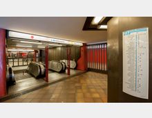 U-Bahnhof mit Rolltreppen zu den Gleisen der Linie U7 Richtung Rathaus Spandau. Links und rechts befinden sich rote Wände, im Vordergrund ein Infopanel mit Linienplan.