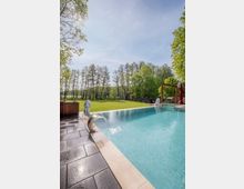 Ein moderner Außenpool mit einer glatten, gefliesten Umrandung und einem kleinen Wasserfall aus Edelstahl, eingebettet in eine grüne Gartenlandschaft mit Rasenflächen und hohen Bäumen im Hintergrund unter klarem Himmel.