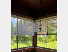 Blick aus einer verglasten Veranda mit Holzrahmen und dunkler Decke, an deren Ecke ein hängender Zieranhänger befestigt ist. An den Fenstern hängen transparente Spitzengardinen; draußen sind eine sonnige Rasenfläche und Bäume zu sehen.