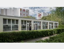 Flaches modernes Gebäude mit großen Fenstern und geöffneten Türen, umgeben von Hecken und Bäumen in einer grünen Umgebung; im Hintergrund befinden sich mehrstöckige Wohngebäude.