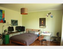 Ein Schlafzimmer mit einem Holzbett, grauer Bettdecke und Kissen, einem Schreibtisch mit zwei Monitoren und einer Lampe sowie einer weiteren Arbeitsfläche mit Stuhl und einigen Büchern. Die Wände sind hellgrün gestrichen und mit Postern und einer Weltkarte dekoriert.