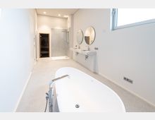 Modernes Badezimmer mit freistehender Badewanne im Vordergrund, Doppelwaschbecken mit zwei runden Spiegeln an der Wand, einer großen Glasdusche und einer Sauna im Hintergrund. Der Raum hat helle Wände, eine minimalistische Gestaltung und ein Fenster auf der rechten Seite.