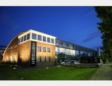 Backsteingebäude mit großen Fenstern und angeschlossenem, modernen Hangar aus Glas und Metall, beleuchtet bei Dämmerung; davor parkende Autos und Grünfläche.