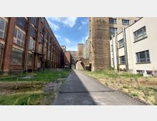 Ein schmaler gepflasterter Weg führt durch einen Innenhof, der von mehrstöckigen, teils roten Backsteingebäuden mit großen Industriefenstern sowie helleren Gebäuden umgeben ist. Der Bereich zeigt Elemente eines alten Fabrikgeländes mit teilweise verwildertem Gras und zurückgebauter Infrastruktur.