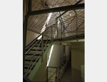 Treppenhaus mit Metalltreppen und Geländern, durchzogen von einem Maschendrahtnetz an der Decke; die Wände sind hell gestrichen, und Lichtstrahlen fallen von oben ins Innere.