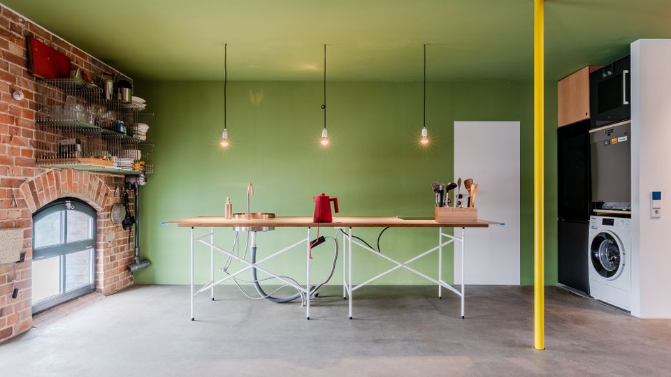 Offene, minimalistische Küche mit grüner Wand, zentraler Kücheninsel aus Holz mit Spüle und drei hängenden Glühbirnenlampen. Links eine freigelegte Ziegelwand mit Bogenfenster und offenen Regalen, rechts ein Geräteschrank mit Waschmaschine sowie eine gelbe Stützsäule auf Betonboden.