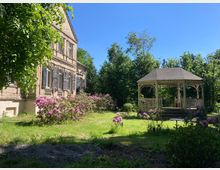 Ein historisches Gebäude mit hellbrauner Fassade und Fensterläden steht links in einem Garten. Rechts daneben befindet sich ein hölzernes Pavillon mit Spitzdach, umgeben von blühenden Rhododendronsträuchern und grünem Rasen. Im Hintergrund sind dichte Bäume zu sehen unter einem klaren, blauen Himmel.