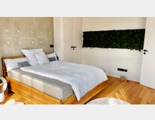 Schlafzimmer mit einem Bett aus Holzrahmen und weißer Bettwäsche, Holzboden und einer vertikalen Mooswand an einer weißen Wand. Im Hintergrund befindet sich eine geschlossene Tür, rechts im Bild ein Ventilator und links eine Steckdose.