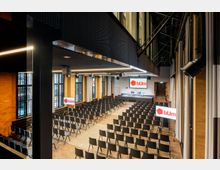 Moderner Konferenzraum mit Backsteinwänden und hohen Fenstern. Der Raum ist mit mehreren Stuhlreihen, einem erhöhten Podium mit zwei Stühlen und Bildschirmen ausgestattet, die das Logo "bUm" zeigen.