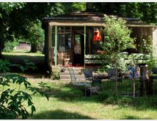 Garten mit einer kleinen, rustikalen Holzhütte, die teilweise von Bäumen umgeben ist. Die Hütte hat eine überdachte Veranda mit Stühlen und Tisch, während der Vorderbereich eine Wiese mit Gartenmöbeln und jungen Bäumen zeigt.
