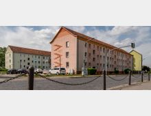 Mehrgeschossige Wohngebäude mit Satteldächern in einem Wohngebiet, umgeben von geparkten Autos und Straßenkreuzungen mit Verkehrsampeln. Im Vordergrund befinden sich Kettenpfosten an einem Gehweg.