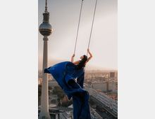Eine Frau in einem langen blauen Kleid schaukelt hoch über einer Stadt, mit dem Berliner Fernsehturm und einer urbanen Skyline bei Sonnenuntergang im Hintergrund.