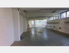 Ein leerer Innenraum eines Industriegebäudes mit weißen Wänden, freiliegenden Rohren und mehreren rechteckigen Fenstern, die Licht hereinlassen. Der Boden besteht aus grauem Beton, und an den Wänden befinden sich Steckdosen und Rohleitungen.