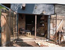 Geöffnete Doppeltüren aus Holz und Glas führen in eine rustikale Werkstatt mit gemauerter Rückwand. Im Innenraum sind Holzstapel, Regale mit Gegenständen und einfache Möbel wie ein Stuhl und ein Hocker zu sehen, während der äußere Bereich mit Ziegelsteinen gepflastert ist.