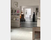 Innenraum einer Kunstgalerie mit weißen Ziegelwänden und grauem Boden, Blick durch einen breiten Rundbogen in einen weiteren Ausstellungsraum. Links stehen Regalflächen mit Büchern und Druckgrafiken, rechts sind gerahmte Bilder an der Wand und eine dunkle Skulptur auf einem Sockel zu sehen.