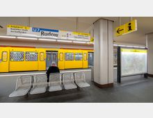Eine U-Bahn-Station in Berlin mit einem gelben U-Bahn-Wagen der Linie U7, der am Bahnsteig steht. Im Vordergrund sind Sitzbänke sowie ein großer Informationsschild zu sehen, und an der Decke hängen Wegweiser mit Ausgängen und Informationen.