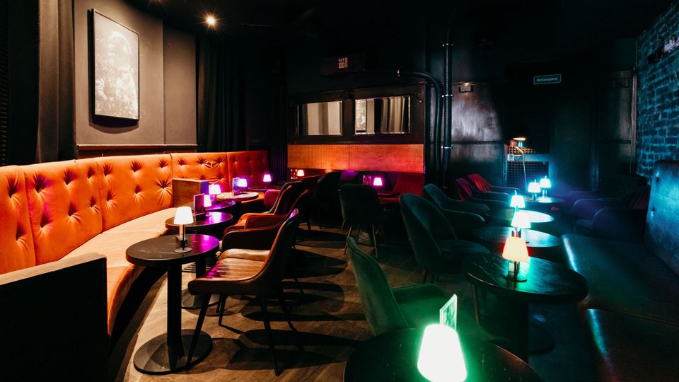 Dunkler Lounge- und Barbereich mit orangefarbener, gepolsterter Sitzbank entlang der Wand, kleinen runden Tischen und Stühlen in Reihen. Auf den Tischen stehen farbige Tischlampen; im Hintergrund sind schwarze Vorhänge, eine Backsteinwand und eine erhöhte Nische mit Sitzplätzen zu sehen.