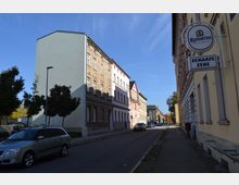 Straßenszene mit mehrstöckigen Wohnhäusern unterschiedlicher Fassadengestaltung auf beiden Seiten, geparkten Autos am Straßenrand und einem Schild mit der Aufschrift „Scharfe Ecke“ rechts im Bild. Klare Herbststimmung mit blauem Himmel und einzelnen Bäumen am Straßenrand.