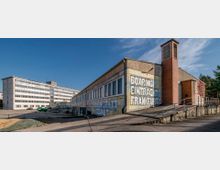 Ein industrielles Gebäude mit großflächigen Graffitis, darunter "Boxring Eintracht Frankfurt", auf einer gelben Wand. Im Hintergrund ein langes, mehrstöckiges Bürogebäude mit weißen Fenstern; vorne eine Rampe und eine rote Backsteinsäule.