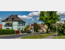 Straßenszene in einem Wohngebiet mit einem mintgrünen, zweistöckigen Haus mit Balkon und braunem Schrägdach auf der linken Seite. Im Hintergrund sind ein Wasserturm, weitere Einfamilienhäuser und ein Straßenschild mit der Aufschrift "O-Metall Weg" zu sehen, umgeben von Bäumen und Hecken.