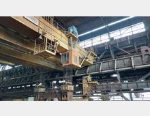 Innenraum einer Industriehalle mit Metallkonstruktionen und einer gelben Brückenkrananlage, die eine orange Steuerkabine und Rohrleitungen trägt. Oberlichter in der Dachkonstruktion lassen Tageslicht einfallen, während der Raum von dunklen Stahlträgern und technischen Elementen dominiert wird.