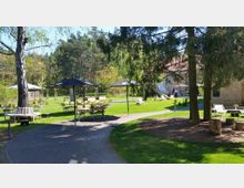 Ein parkähnlicher Außenbereich mit grüner Wiese, Bäumen und Sitzgelegenheiten aus Holz unter Sonnenschirmen. Ein geschotterter Weg führt vorbei an einer Terrasse mit Liegestühlen und einem Gebäude mit hellem Mauerwerk im Hintergrund.