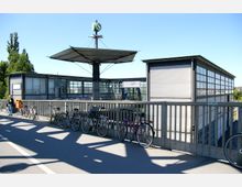 Moderne S-Bahn-Station mit einem flachen Dach und seitlichen Glas- und Metallwänden. Im Vordergrund eine Brücke mit abgestellten Fahrrädern; im Hintergrund Bäume und ein klarer Himmel.