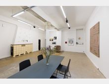 Heller Innenraum einer Galerie oder eines Ateliers mit weiß gestrichenen Wänden, moderner Kunst an den Wänden und einer minimalistischen Einrichtung. Im Vordergrund steht ein dunkler Tisch mit schwarzen Stühlen und einer Vase mit Blumen, im Hintergrund ein Sessel, ein Kachelofen und industrielle Deckenbeleuchtung.