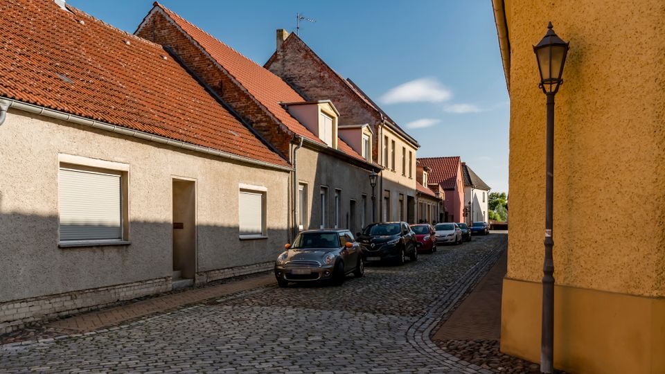 Schmale Kopfsteinpflasterstraße in einem Wohnviertel mit niedrigem Reihenhausbestand und roten Ziegeldächern. Entlang der Straße stehen mehrere geparkte Autos; an der rechten Hauswand befindet sich eine Straßenlaterne.