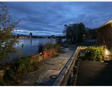 Uferpromenade an einem ruhigen Fluss bei Dämmerung, mit betoniertem Weg, Geländer und einer beleuchteten Holzterrasse neben einem Gebäude. Im Hintergrund spiegeln sich Lichter einer Stadt skyline im Wasser unter dicht bewölktem Himmel, flankiert von Bäumen und Ufervegetation.