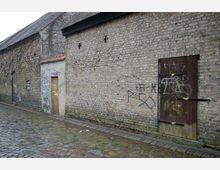 Alte Backsteinfassade mit verwitterten Türen und Graffiti, entlang einer gepflasterten Straße. Die Wand ist teilweise mit rankenden Pflanzen bewachsen.