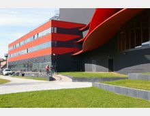 Modernes Gebäude mit einer auffälligen roten und schwarzen Fassade, großflächigen Fenstern und einer geschwungenen Dachkonstruktion. Vor dem Gebäude befinden sich ein gepflasterter Weg, grüne Rasenflächen und mehrere Personen.